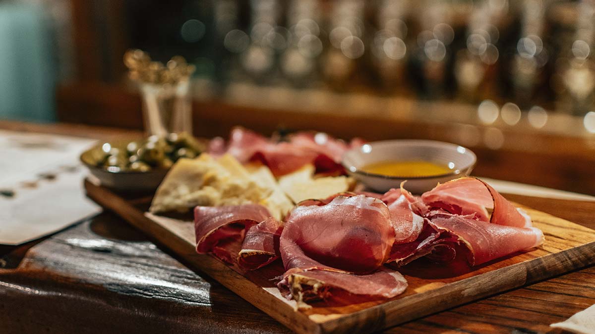 Charcuterie board ideas featuring sliced prosciutto, cheese, olives, and honey on a wooden serving board