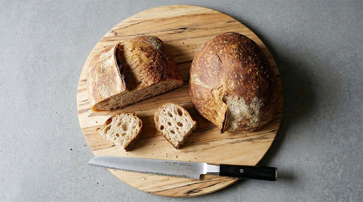 Rustic sourdough bread sliced on a round wooden board with serrated knife
