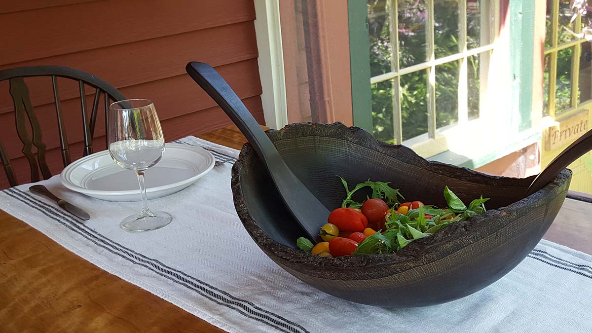 Ebonized live edge wooden salad bowl with serving utensils on a set dining table