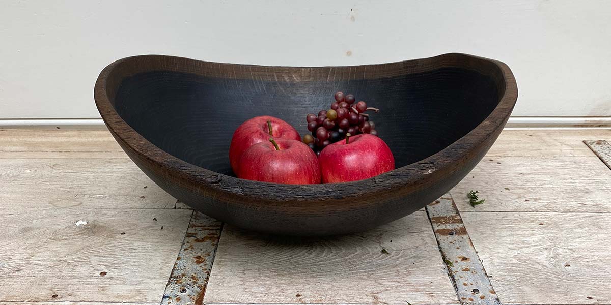 Black ebonized wooden bowl holding red apples and grapes on a rustic surface