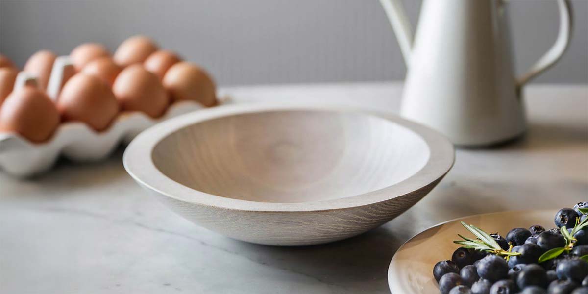 Light-toned white oak wooden bowl on a kitchen counter with eggs and fresh ingredients nearby